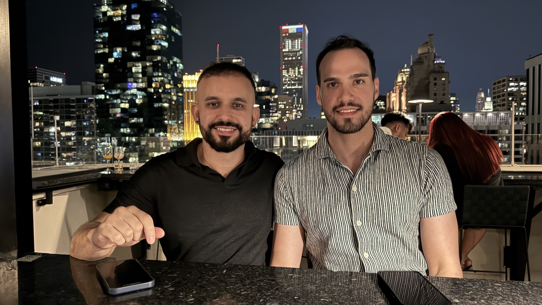 Leo and Carlos on a rooftop at night, city skyline behind them